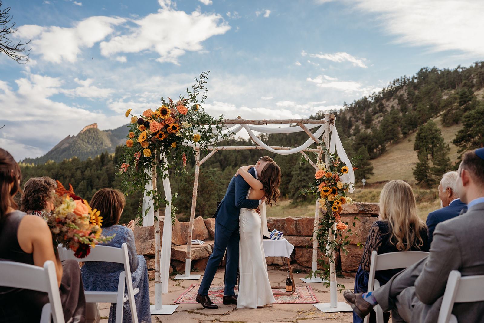 The bride and groom kissing beneath a floral ceremony arch with mountain views, surrounded by seated guests during their wedding ceremony.