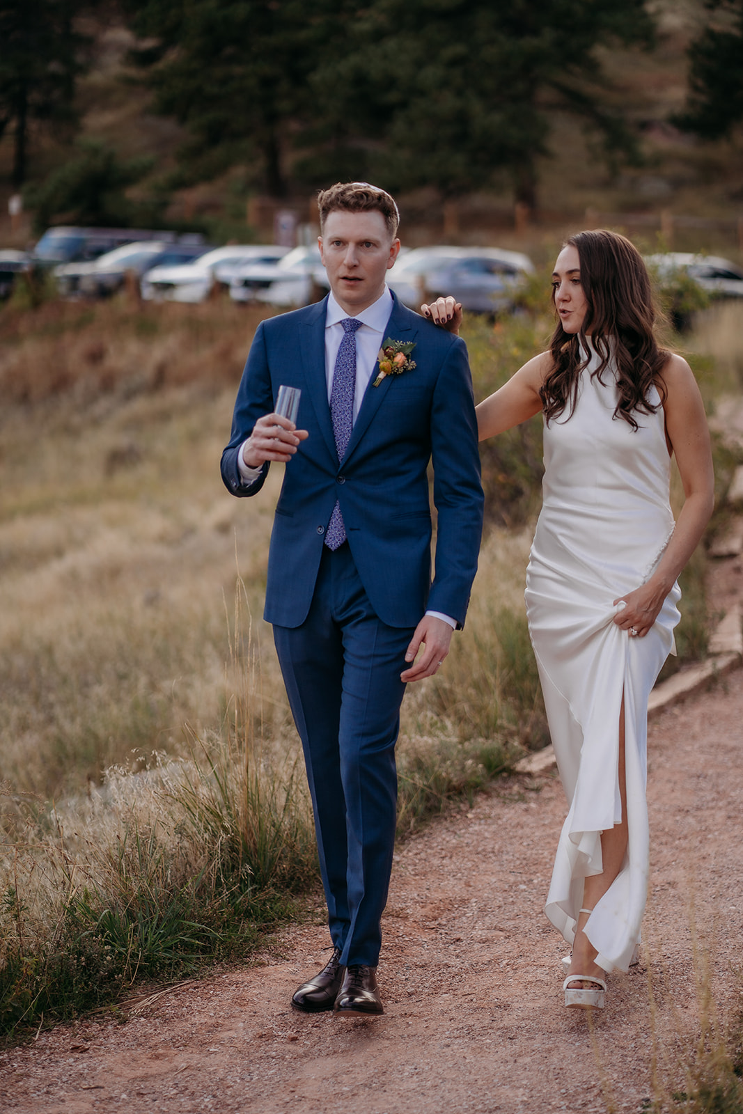 A bride and groom walking along a dirt path, the groom holding a drink while talking together during a relaxed Colorado wedding moment.