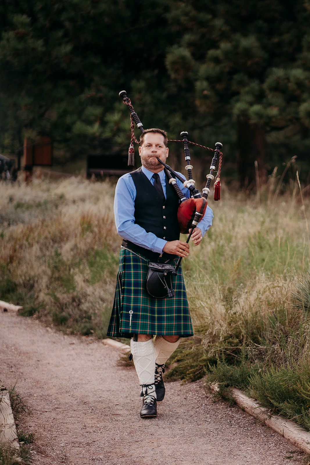 A bagpiper wearing a traditional kilt walking along a mountain path while playing music during a Colorado wedding ceremony.