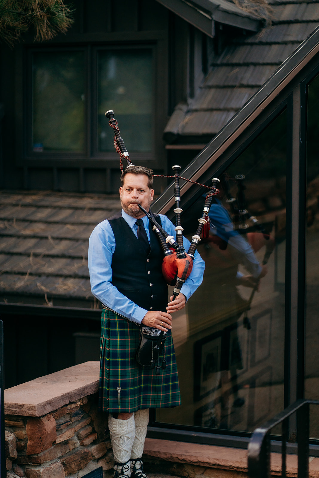 A bagpiper standing and playing music near a stone building, adding live ceremony music to a Colorado wedding celebration.