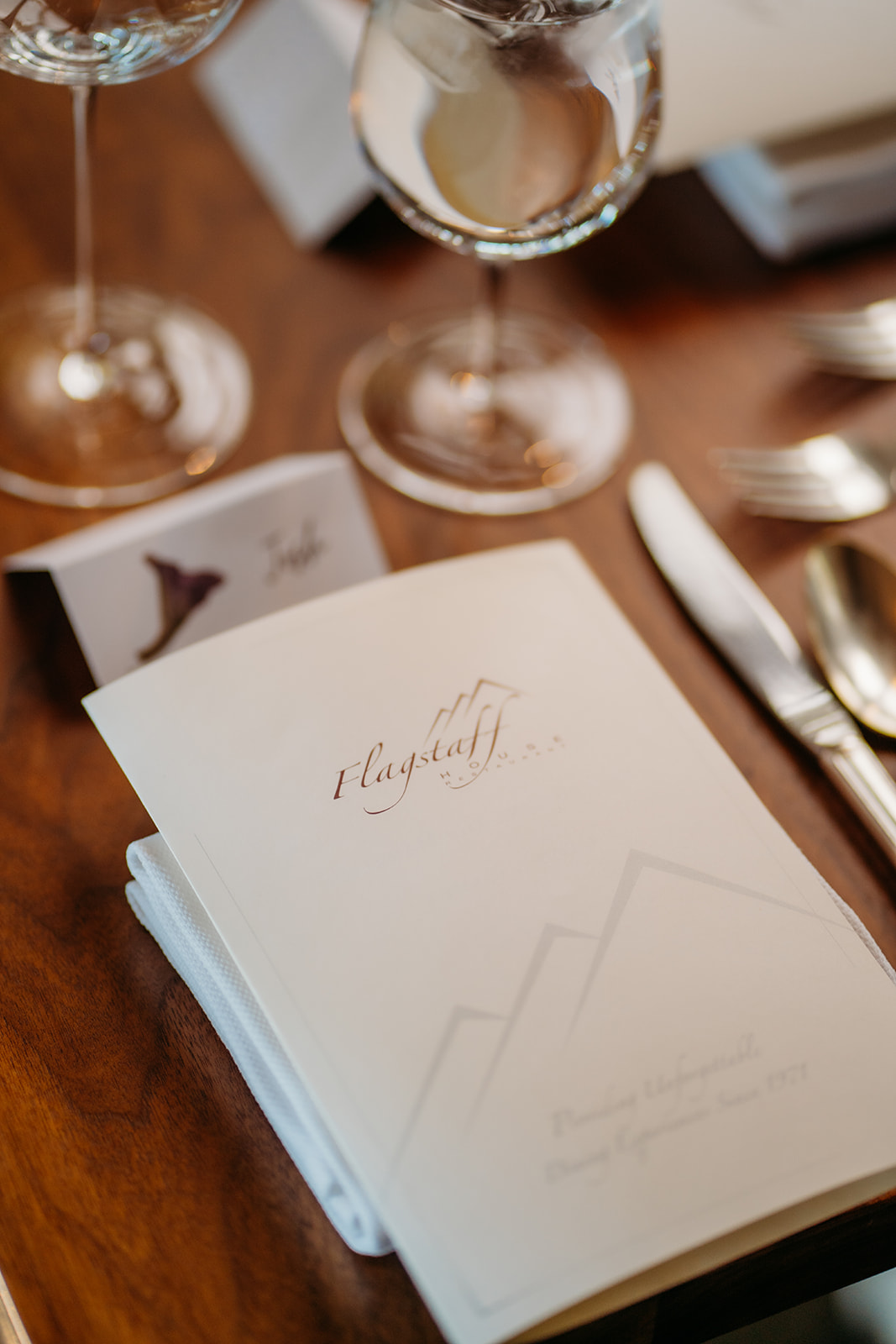 A close-up of elegant reception place settings with menus, glassware, and folded napkins arranged on a wooden table for a wedding dinner.