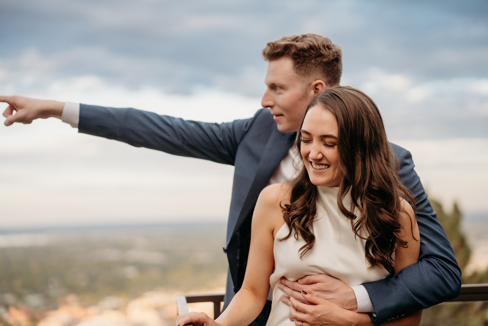A groom pointing out toward the view while standing behind his bride on a deck overlooking the landscape, captured during Colorado weddings.