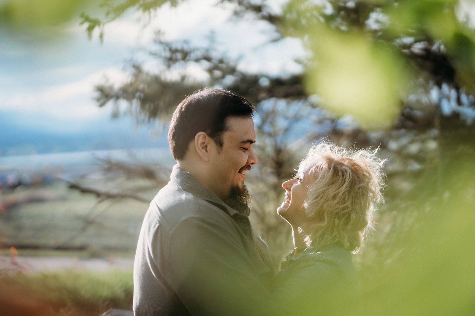 A joyful, candid moment between a couple laughing together under a tree, with the Rockies in the distance—captured by a Denver engagement photographer.