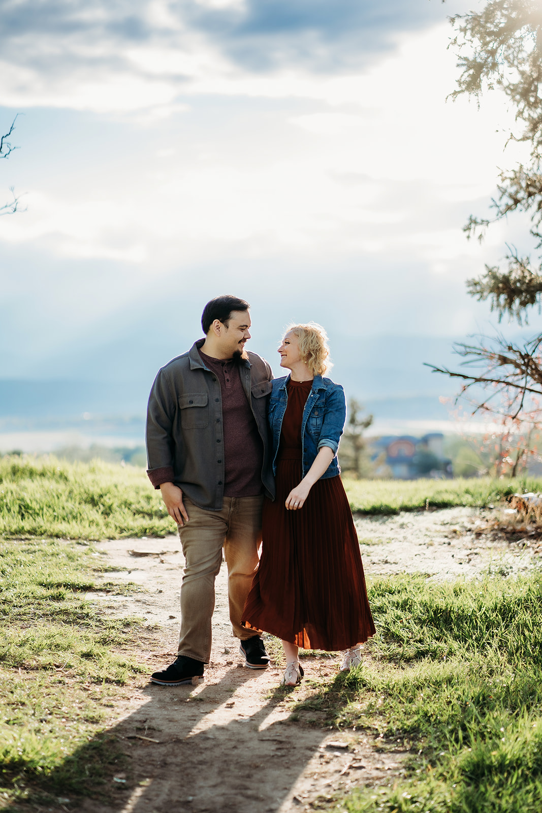 A couple walks hand in hand along a dirt trail with mountain views and glowing light—captured by a Denver engagement photographer.