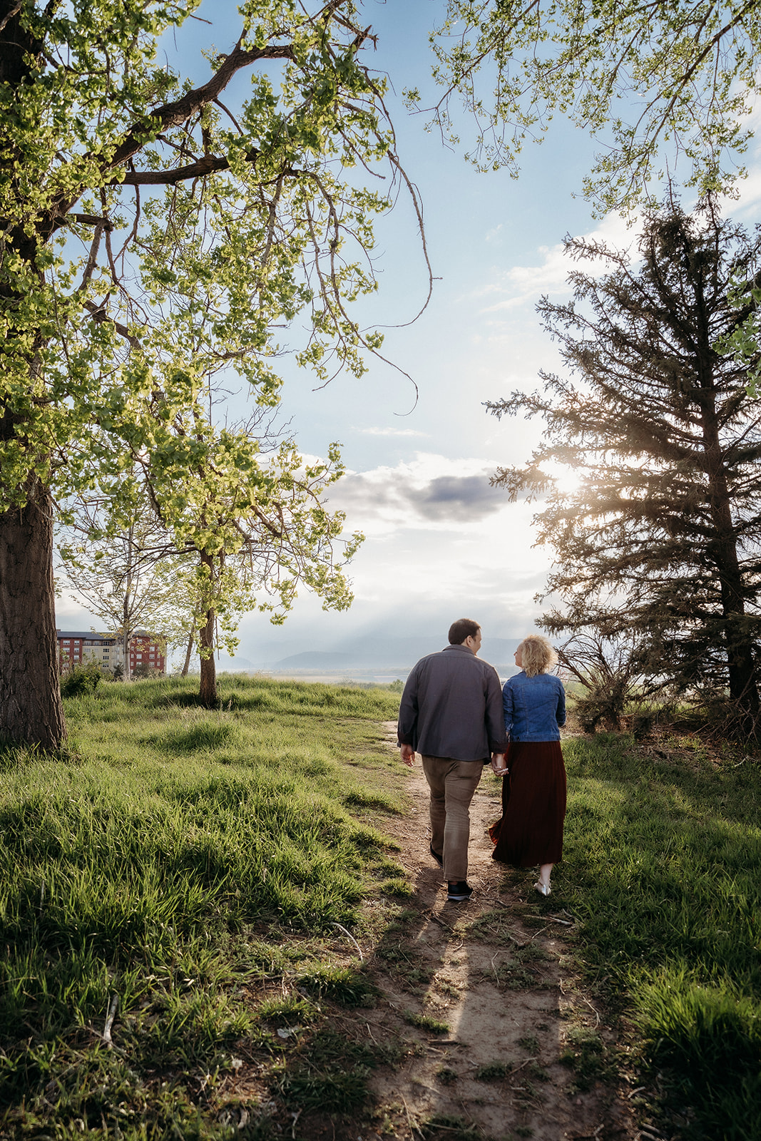 A couple walks hand in hand along a tree-lined trail, surrounded by spring greenery and soft evening light.