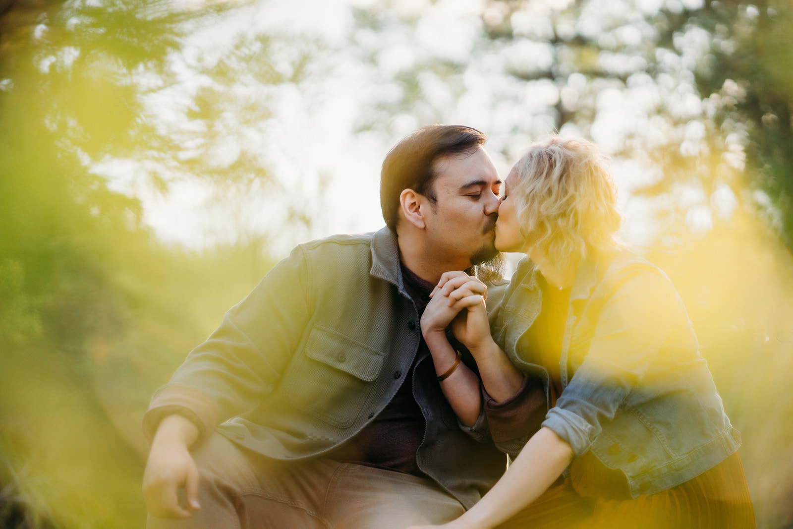 A candid kiss through soft yellow foreground leaves, as the couple holds hands and sits nestled in nature.