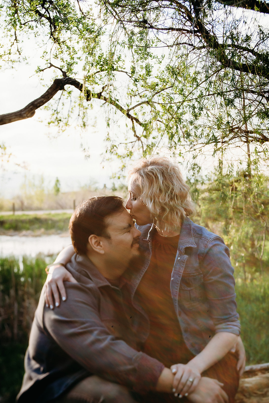 A woman gently kisses her partner’s forehead beneath a tree, with sunlight and a quiet creek behind them—Denver engagement photographer.