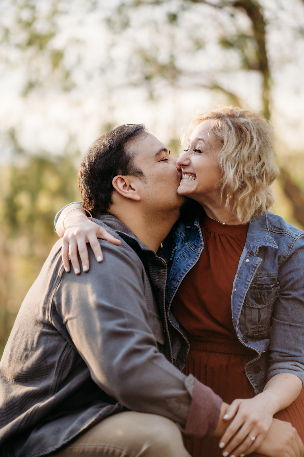 A joyful nose kiss shared between a couple sitting close in the woods, surrounded by warm afternoon light.