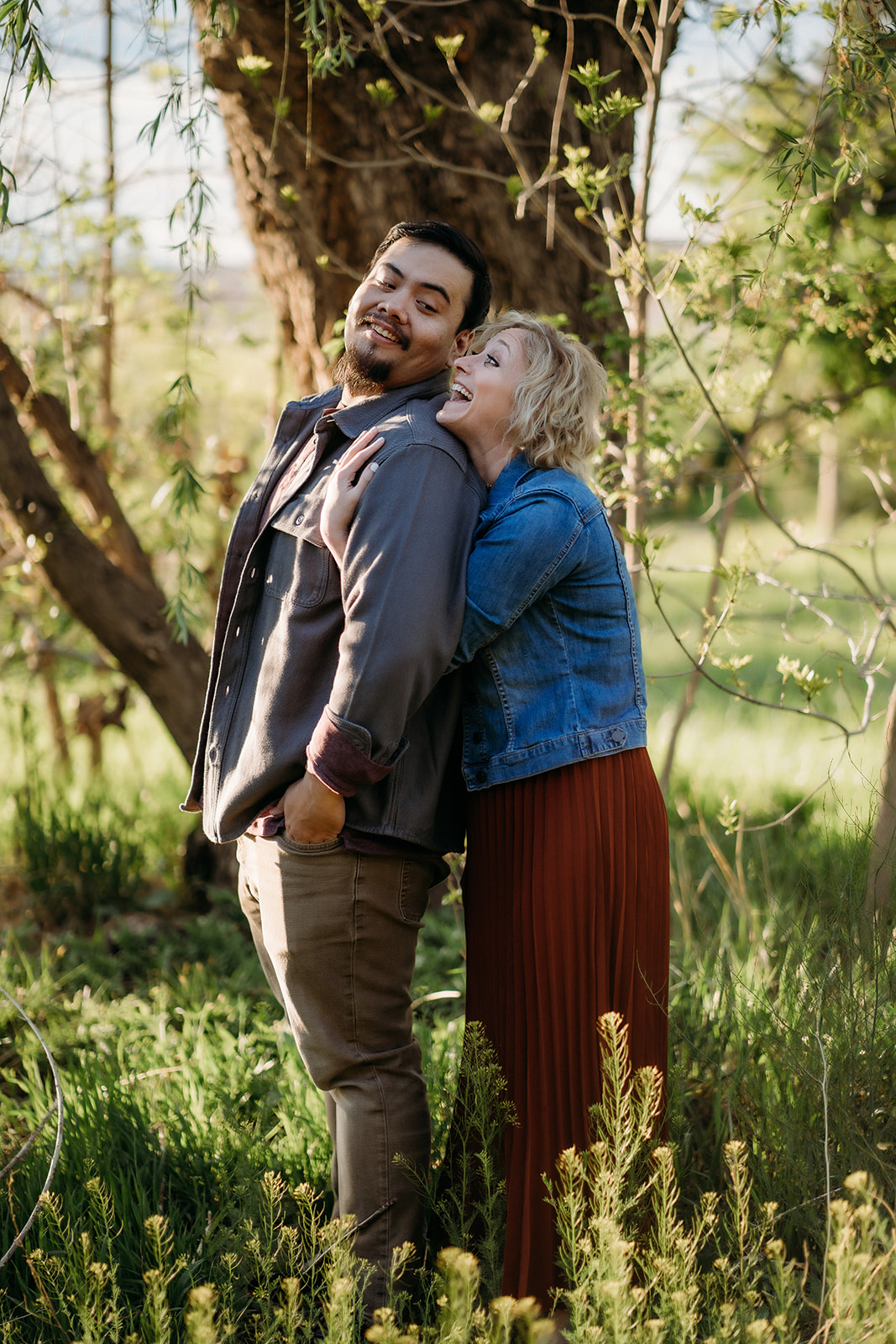 A lighthearted pose as a woman wraps her arms around her partner from behind, laughing under the trees—Denver engagement photographer.