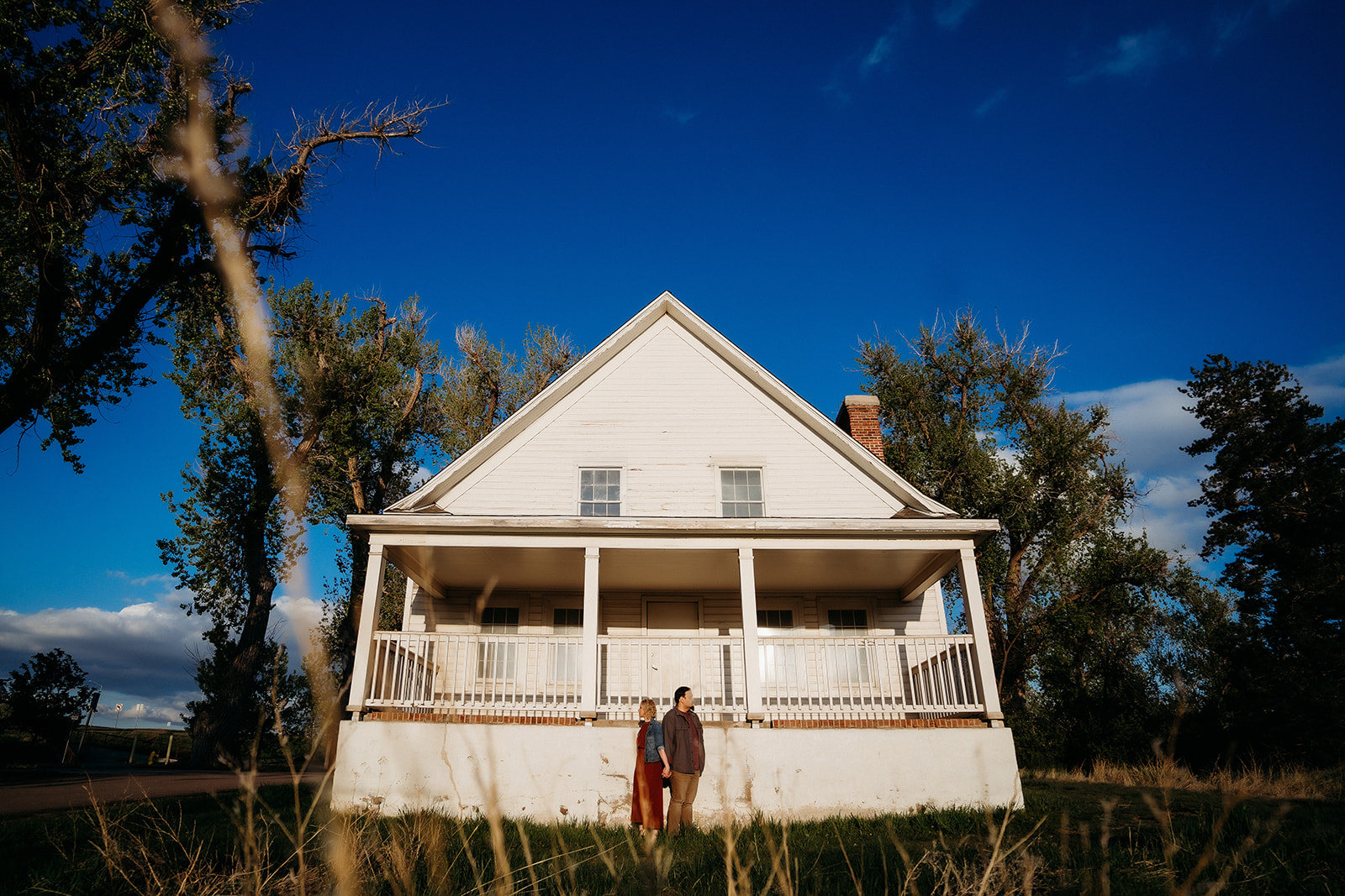 Couple stands in front of a historic white farmhouse, holding hands beneath a bright blue sky in rural Colorado.