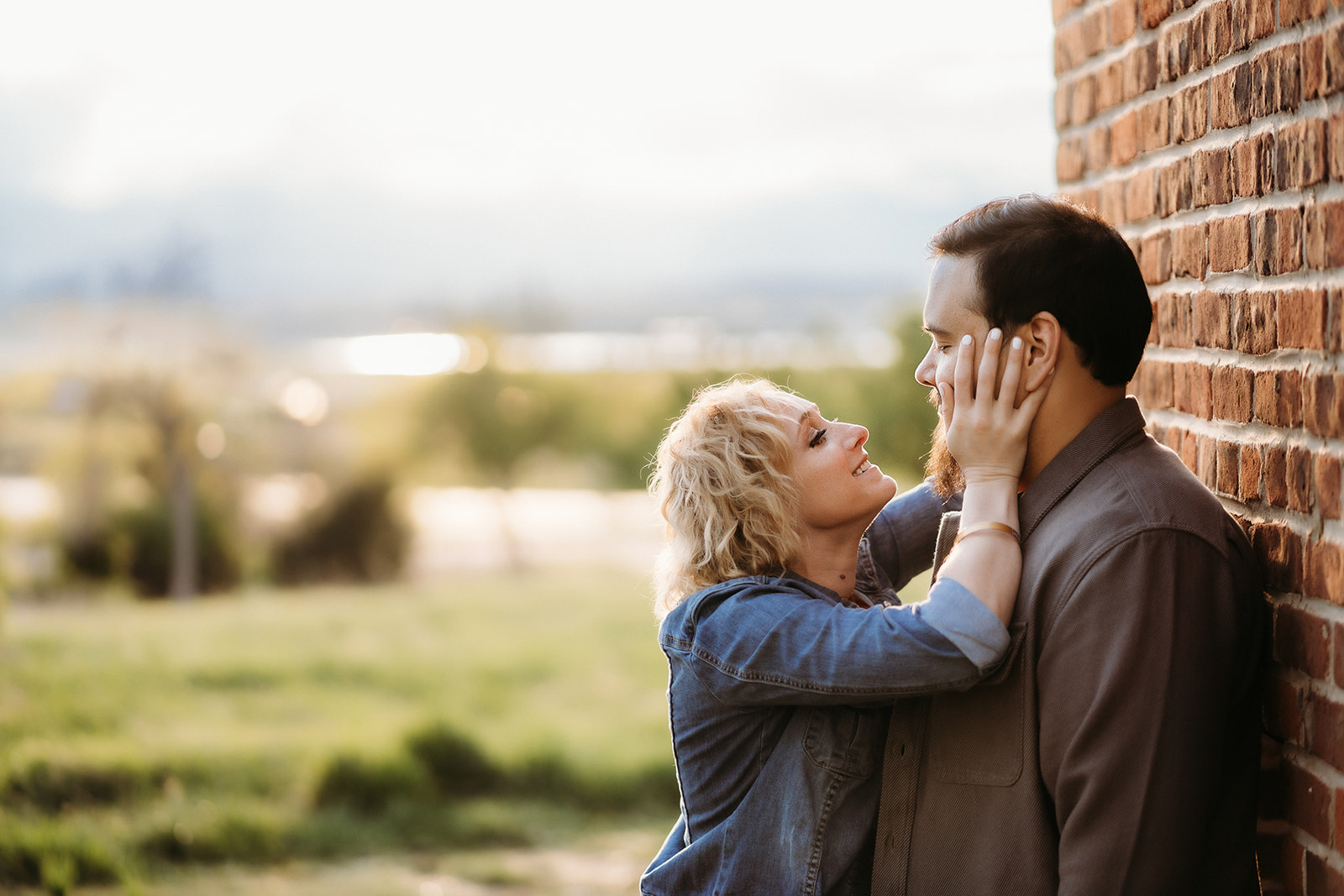 A couple shares a sweet moment against a brick wall, with golden-hour light and mountain views in the background.