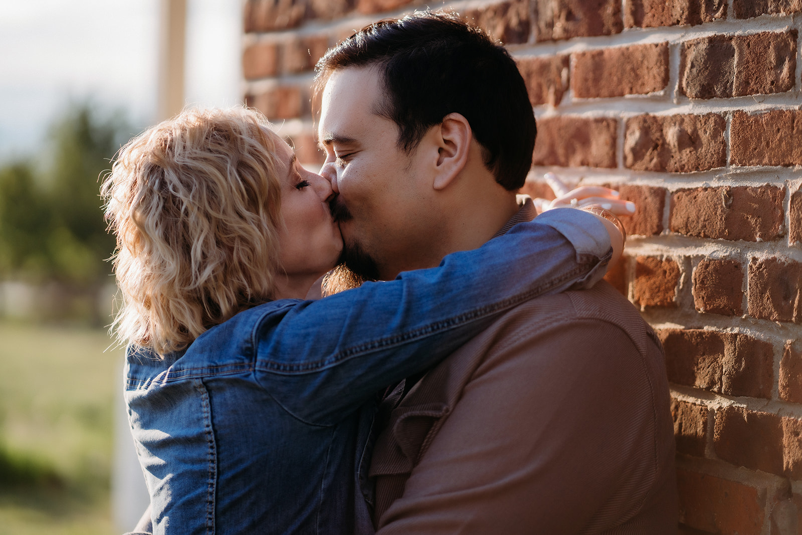 A playful kiss against a red brick wall, with the couple smiling through the moment, wrapped in each other's arms.