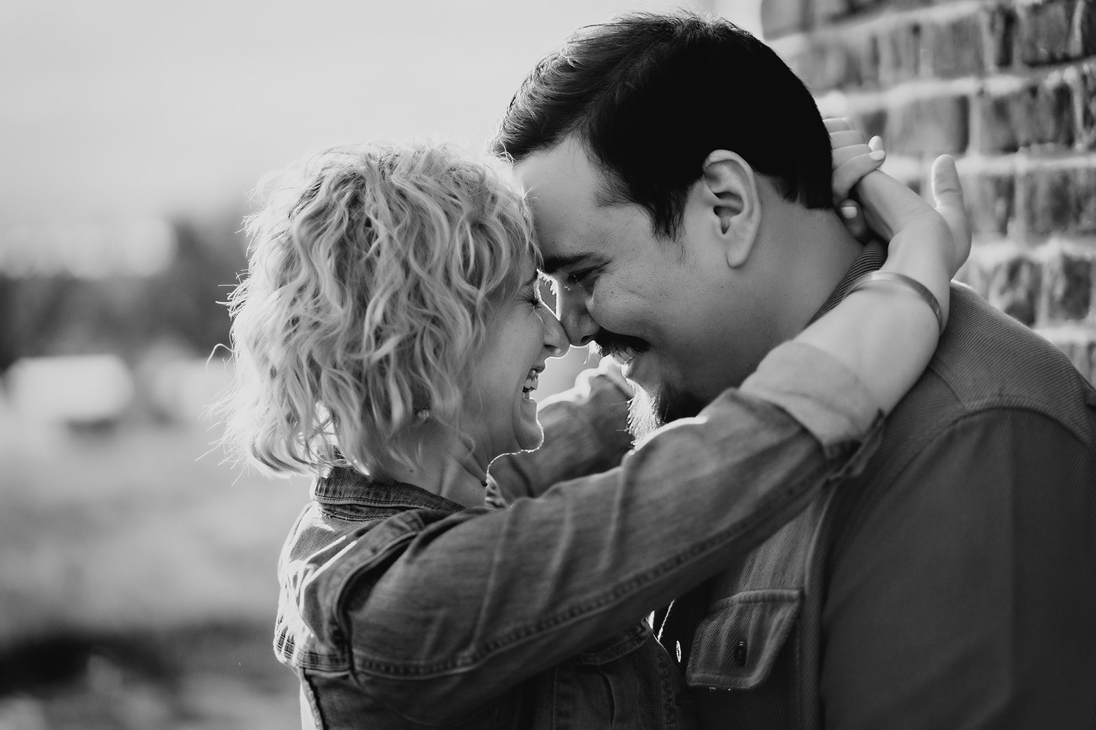 A black and white photo of engaged couple touching noses against brick wall laughing together captured by denver engagement photographer.