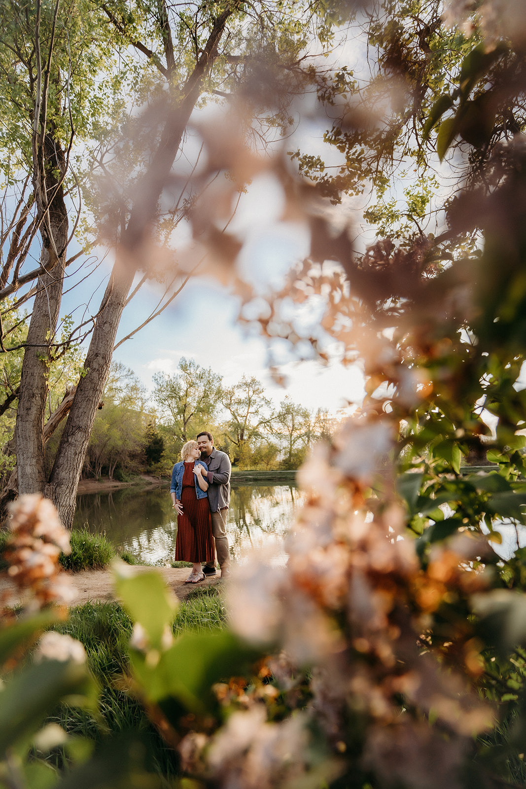 Artistic photo of a couple by a lake, framed through blooming branches, evoking springtime romance in Colorado.