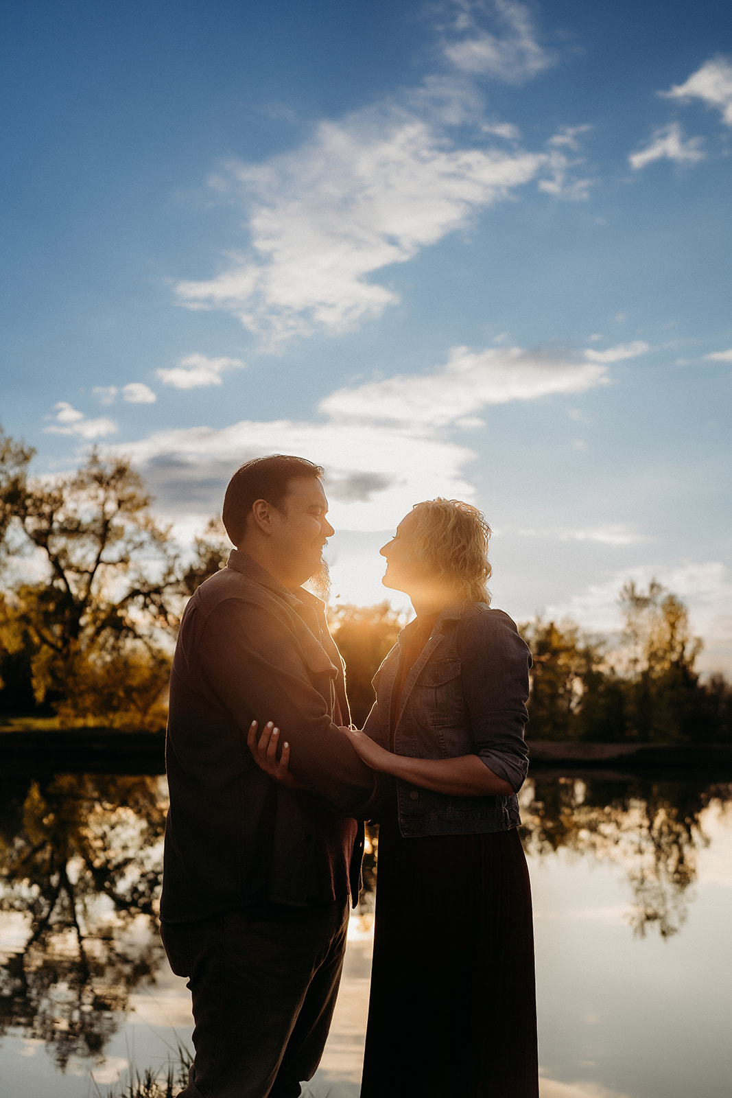 Silhouetted against a glowing sky, the couple stands by a reflective lake, wrapped in golden hour light.