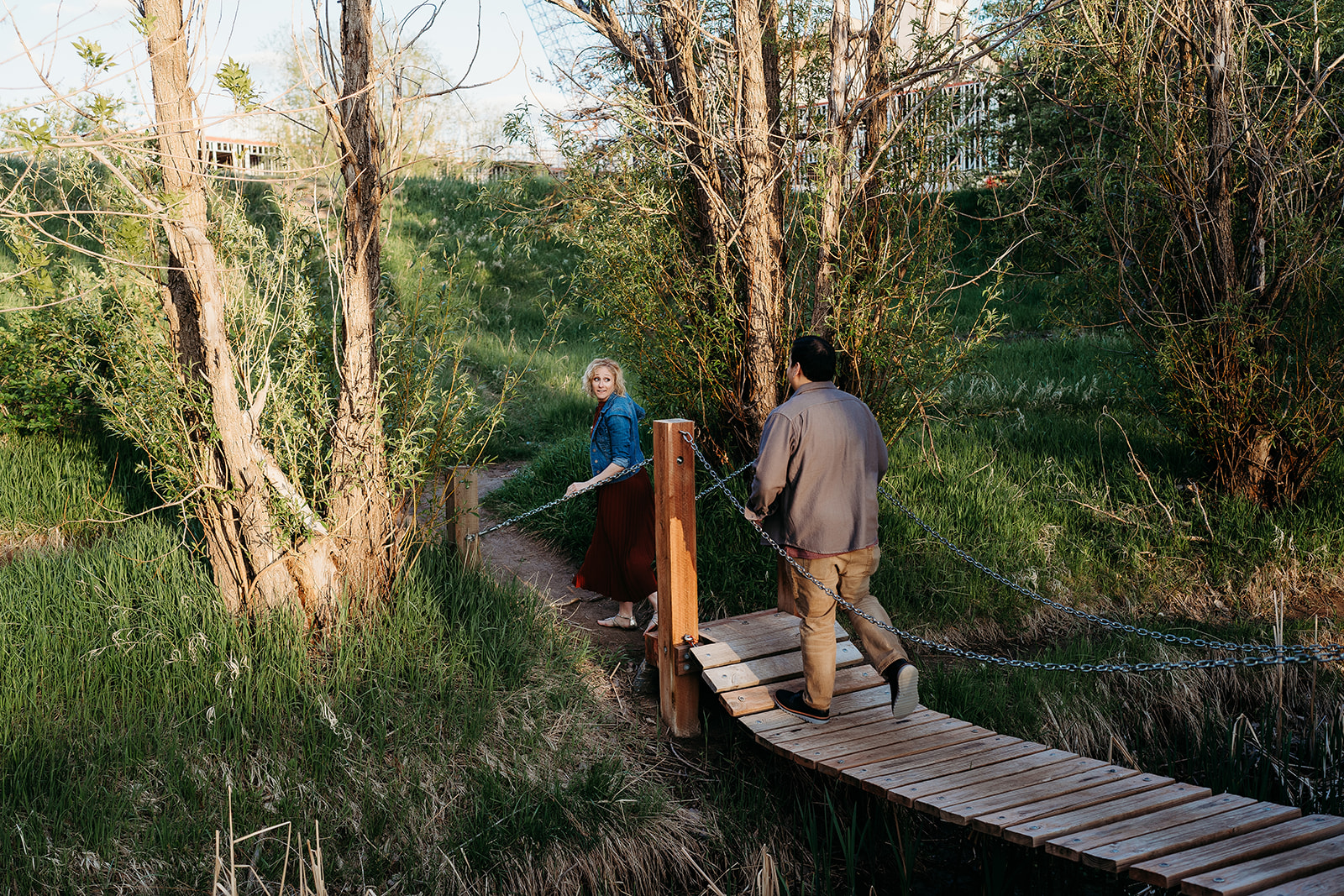A couple walks across a wooden footbridge surrounded by tall grass and trees, enjoying a peaceful spring evening.
