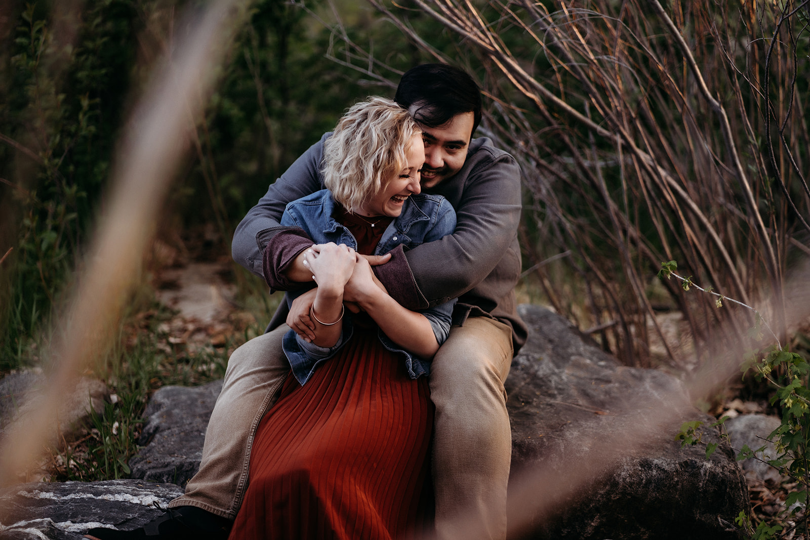Cozy and candid, the couple laughs while cuddling on a boulder surrounded by tall grasses and wild branches.