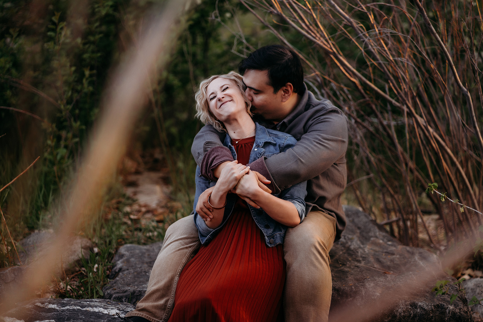 Wrapped in a playful embrace, the couple shares a laugh while sitting on a boulder in the woods—captured by a Denver engagement photographer.