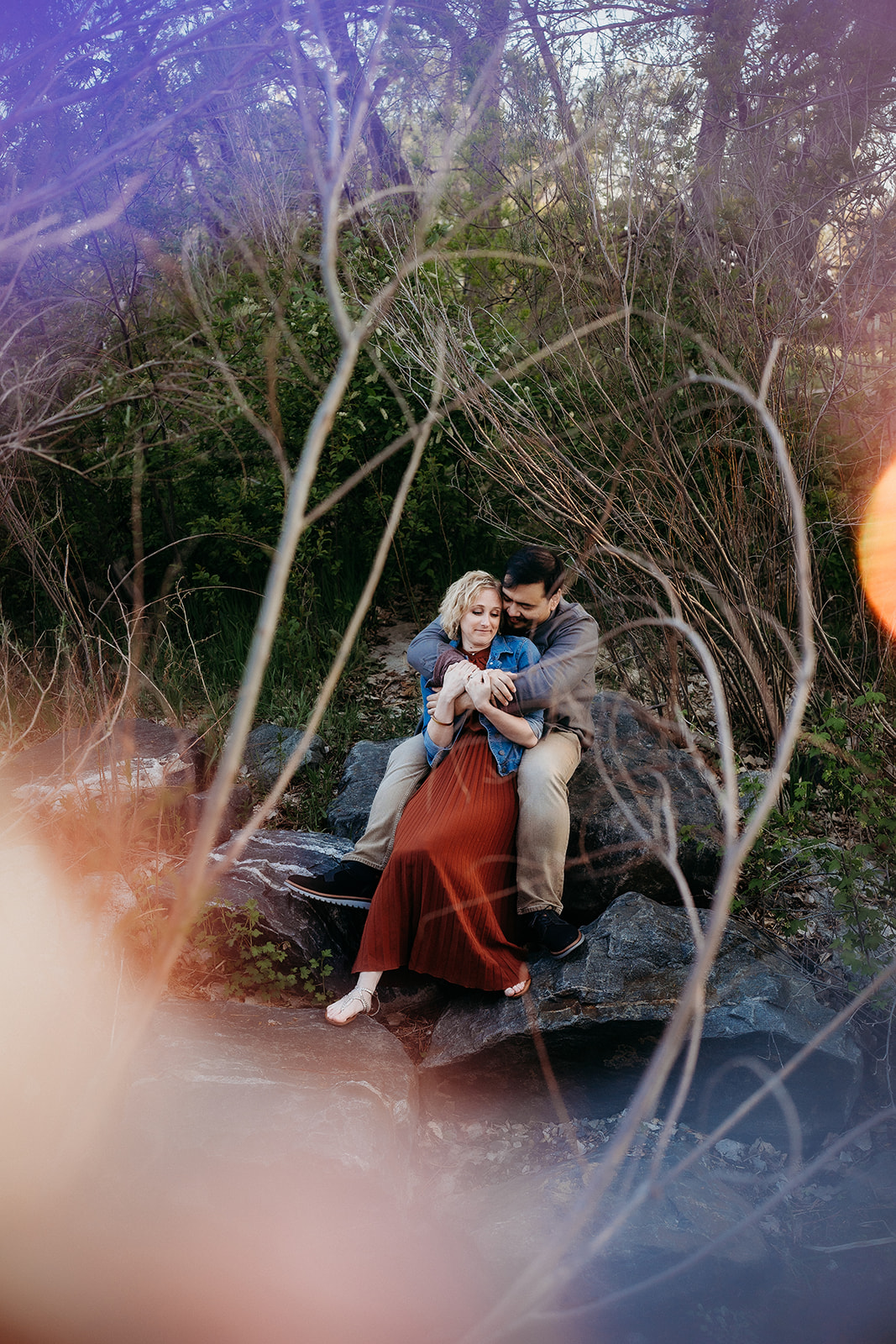 Artistic lens flare surrounds a cozy couple cuddling on a boulder in the woods, full of laughter and connection.