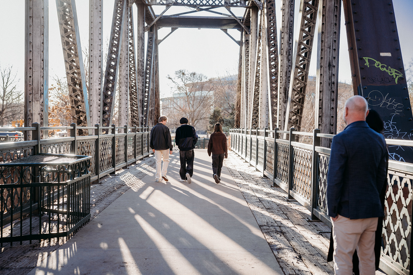 Three siblings walking away together across a historic Denver bridge as a parent watches, captured candidly by Denver photographers.
