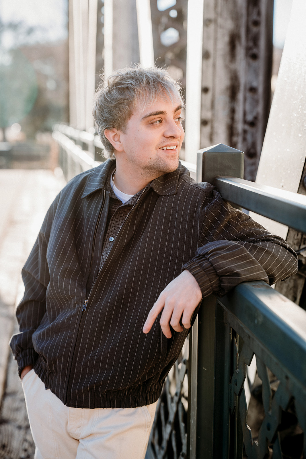 A young man leaning casually on a bridge railing in Denver, smiling softly during an urban lifestyle portrait session.