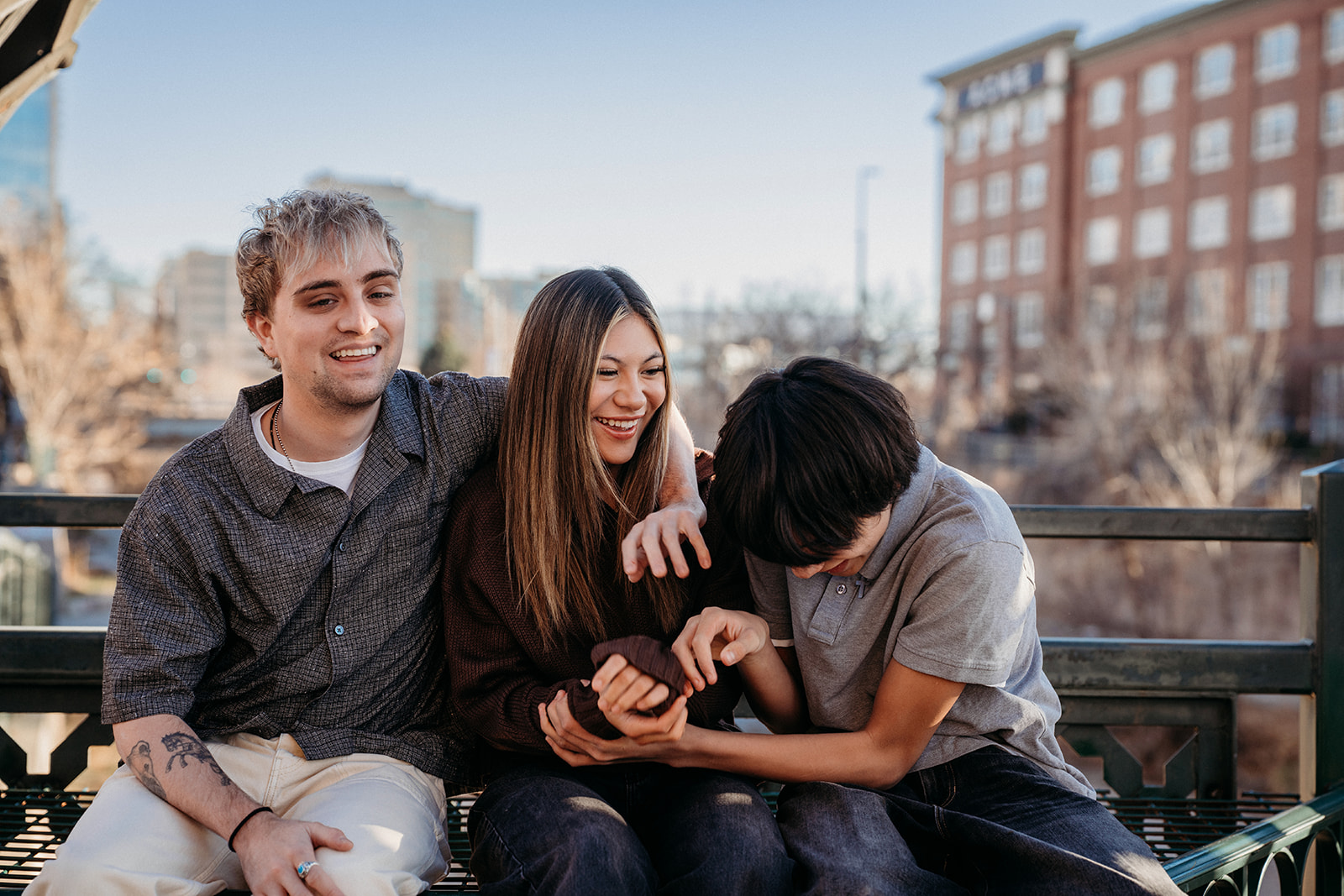 Three siblings sitting together on a bench in downtown Denver, laughing and connecting during a relaxed lifestyle family photography session.