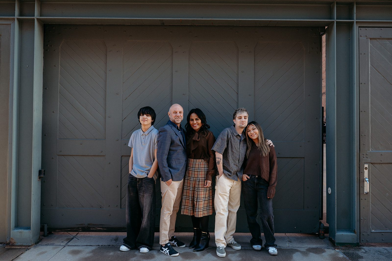 A family of five standing shoulder to shoulder in front of dark industrial doors, smiling naturally during a downtown Denver family session.