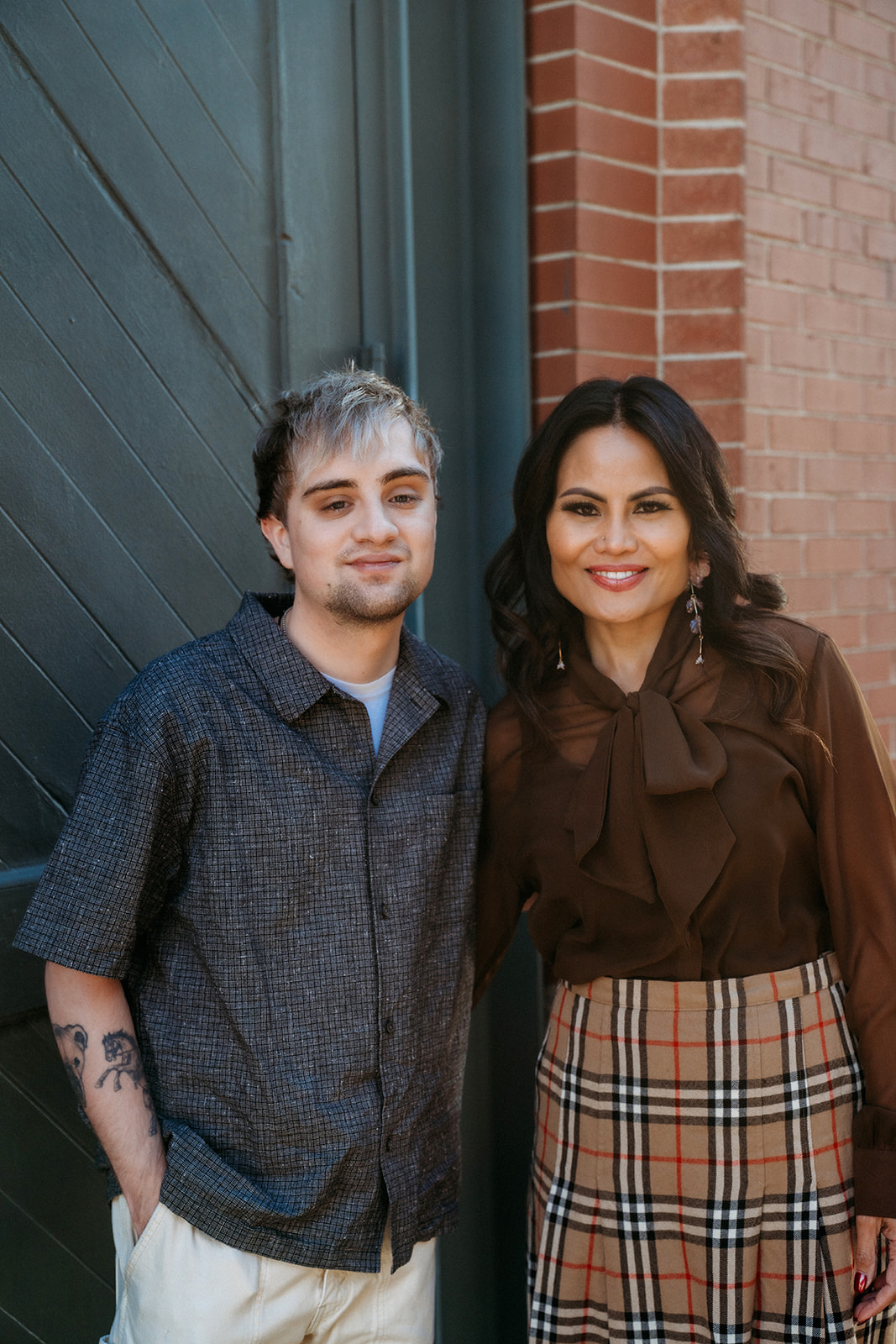A mother standing beside her adult son against a brick building in Denver, photographed in a warm, natural light family session.
