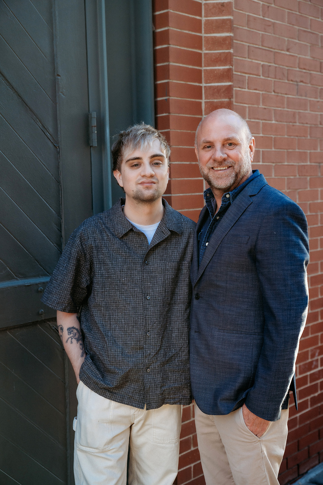 A father and adult son standing side by side against a brick building in Denver, smiling naturally during a family portrait session.