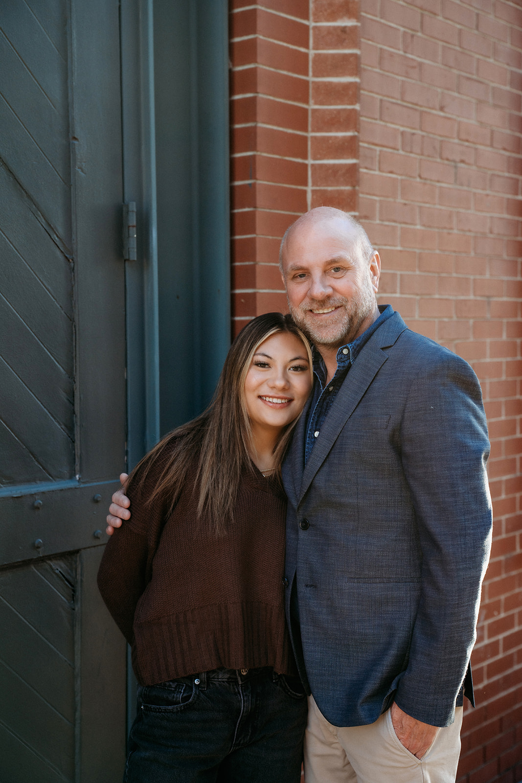 A father and daughter standing close together against a brick wall in Denver, smiling during a relaxed family photography session.