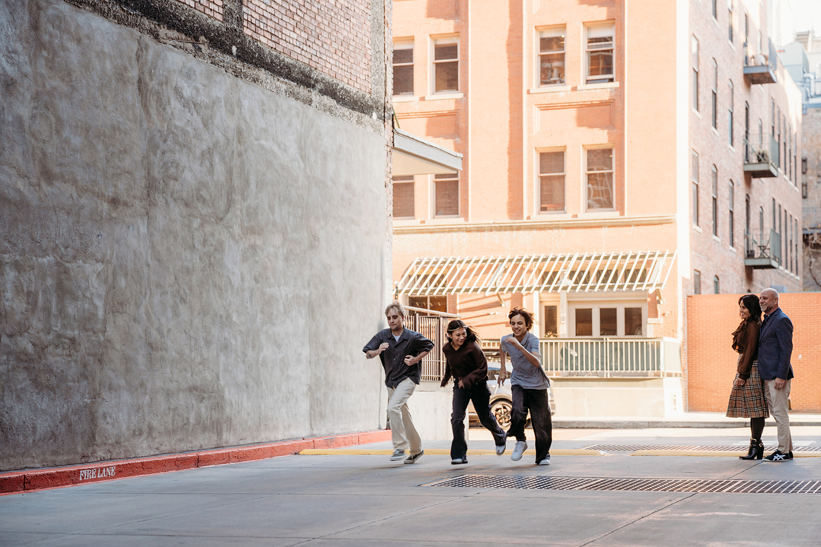 Three siblings running and laughing through a downtown Denver alleyway while parents watch from the background during a candid family session.