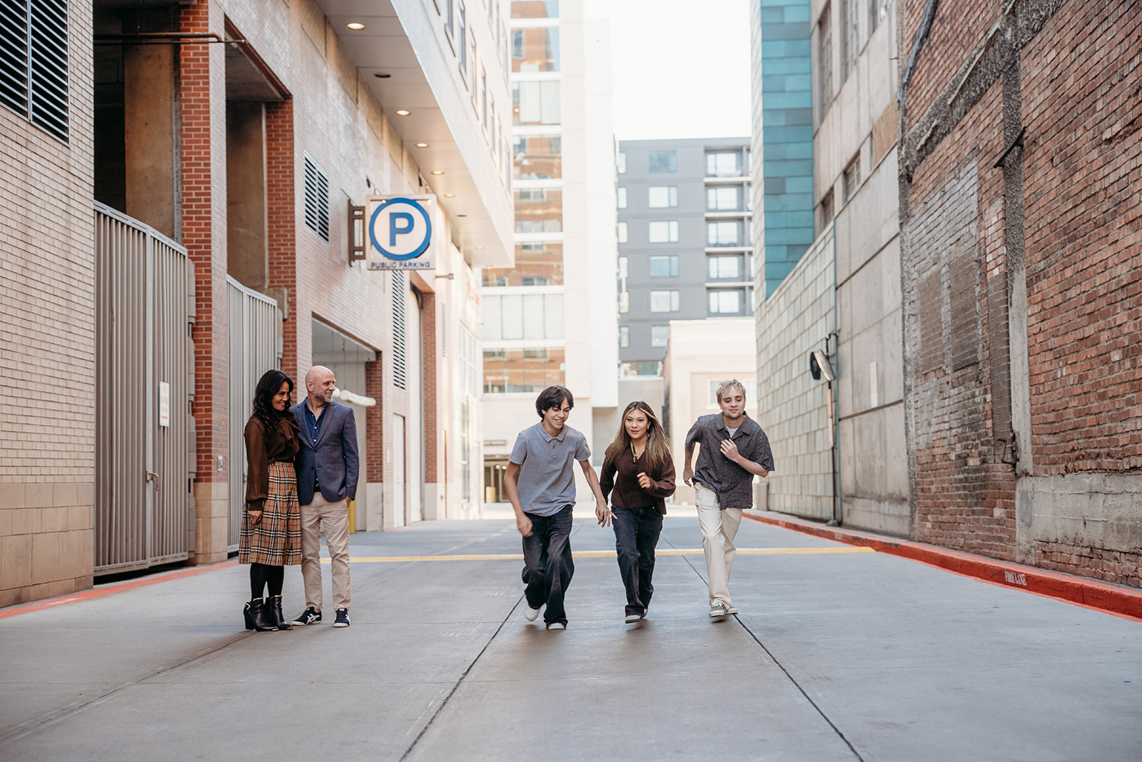 Three siblings running and laughing down a downtown Denver alley while their parents walk behind them, captured candidly by Denver photographers during a family session.
